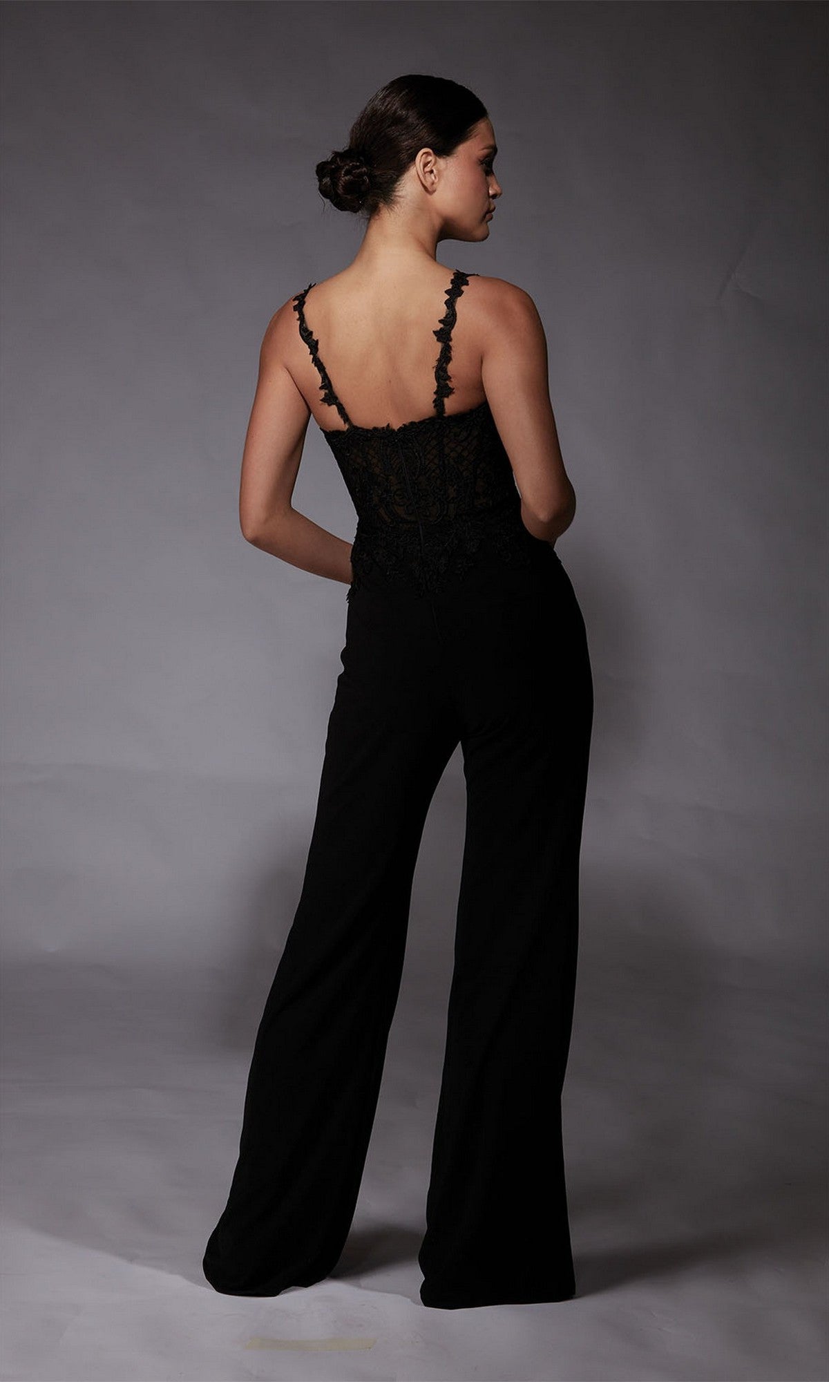 A woman faces away, wearing the Alyce 70074—a long formal black lace jumpsuit with spaghetti straps and high-waisted wide-leg pants—against a plain gray backdrop.